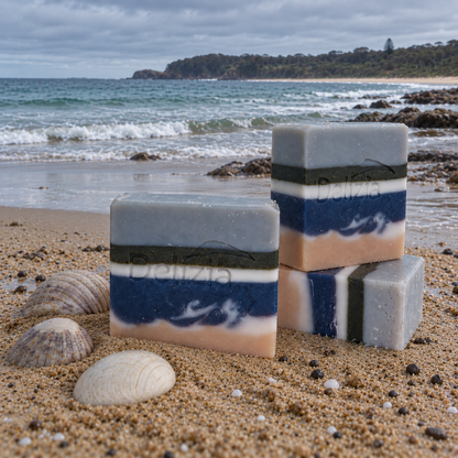 Three Delizia Naturally Aussie Summer soap bars with blue, white and sandy layers beside seashells on a beach