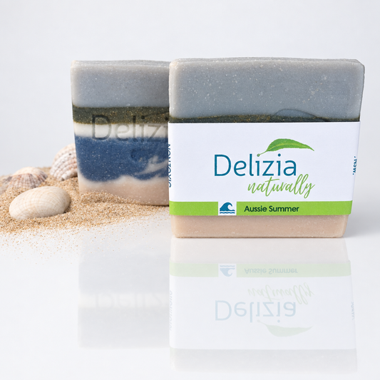 Two Delizia Naturally Aussie Summer soap bars, one labelled and one showing its stamped logo and ocean-inspired layers, beside sand and shells on a white reflective surface