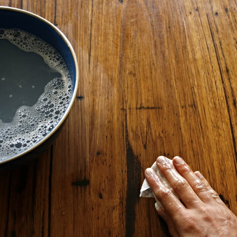 Hand wiping a timber surface beside a bowl of foamy liquid